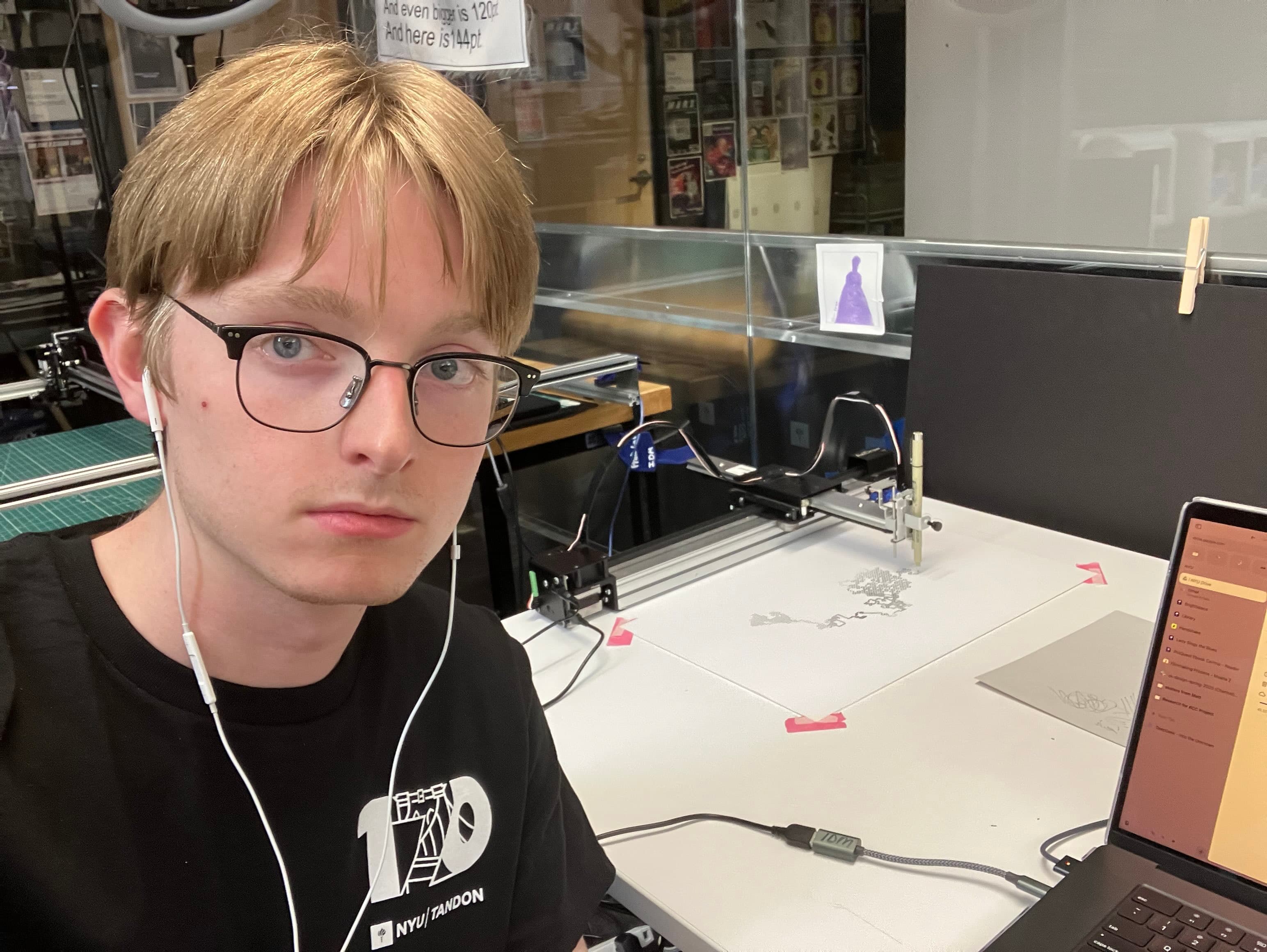 A man taking a selfie at a desk. To his left, a plotter printer is printing black dots on white paper